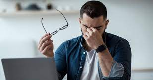 A man experiencing eye strain, holding his glasses in one hand and rubbing his eyes, seated in front of a laptop.